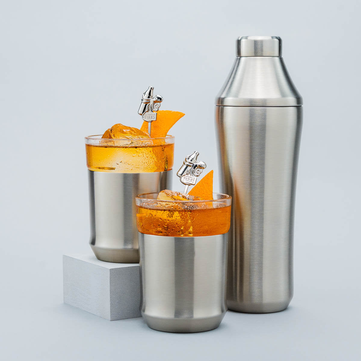 Two cocktail glasses with orange drinks and a metal shaker on a light gray background