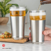Man pouring cold beer from a bottle into the Elevated Craft Hybrid Pint Glass in brushed stainless finish next to another stainless hybrid pint glass full of cold beer with the 2024 red dot design award badge in the bottom left corner of image
