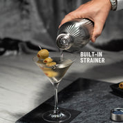Hand pouring liquid through strainer of Stainless Steel Elevated Craft Diamond Edition Hybrid Cocktail Shaker into martini glass with olives
