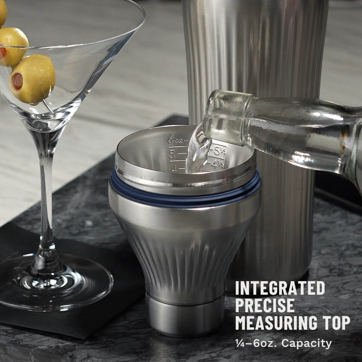 Bottle pouring clear liquid into Stainless Steel Elevated Craft Diamond Edition Hybrid Cocktail Shaker measuring top next to tumbler and martini glass with olives
