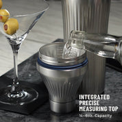 Bottle pouring clear liquid into Stainless Steel Elevated Craft Diamond Edition Hybrid Cocktail Shaker measuring top next to tumbler and martini glass with olives