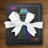 A boxed product wrapped in a large white ribbon bow, photographed from above on a wooden surface. The box is dark with a colorful neon-style bottle illustration and the words 'ELEVATED ART' visible near the top.