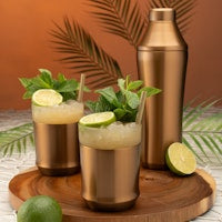 Three copper-colored cocktail mugs filled with a pale yellow drink and lime slices, alongside a matching copper shaker and a tall copper tumbler, arranged on a wooden tray with limes and tropical leaves in the background.