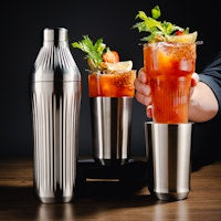 A stainless steel cocktail shaker set shown with two filled glasses of iced drink garnished with mint, on a dark wooden table with a person's hand holding one glass.