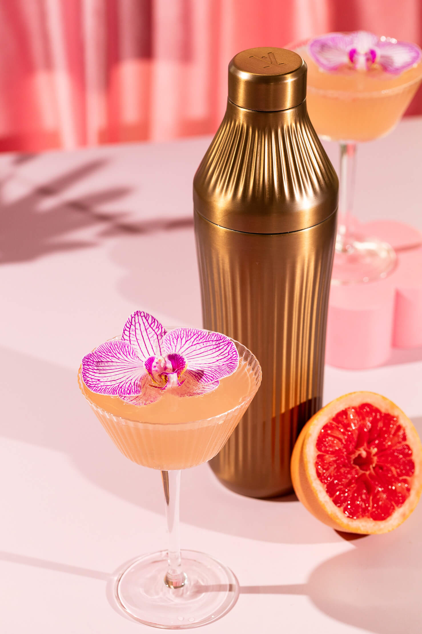 Cocktail with pink orchid garnish, gold shaker, and grapefruit on a pink background