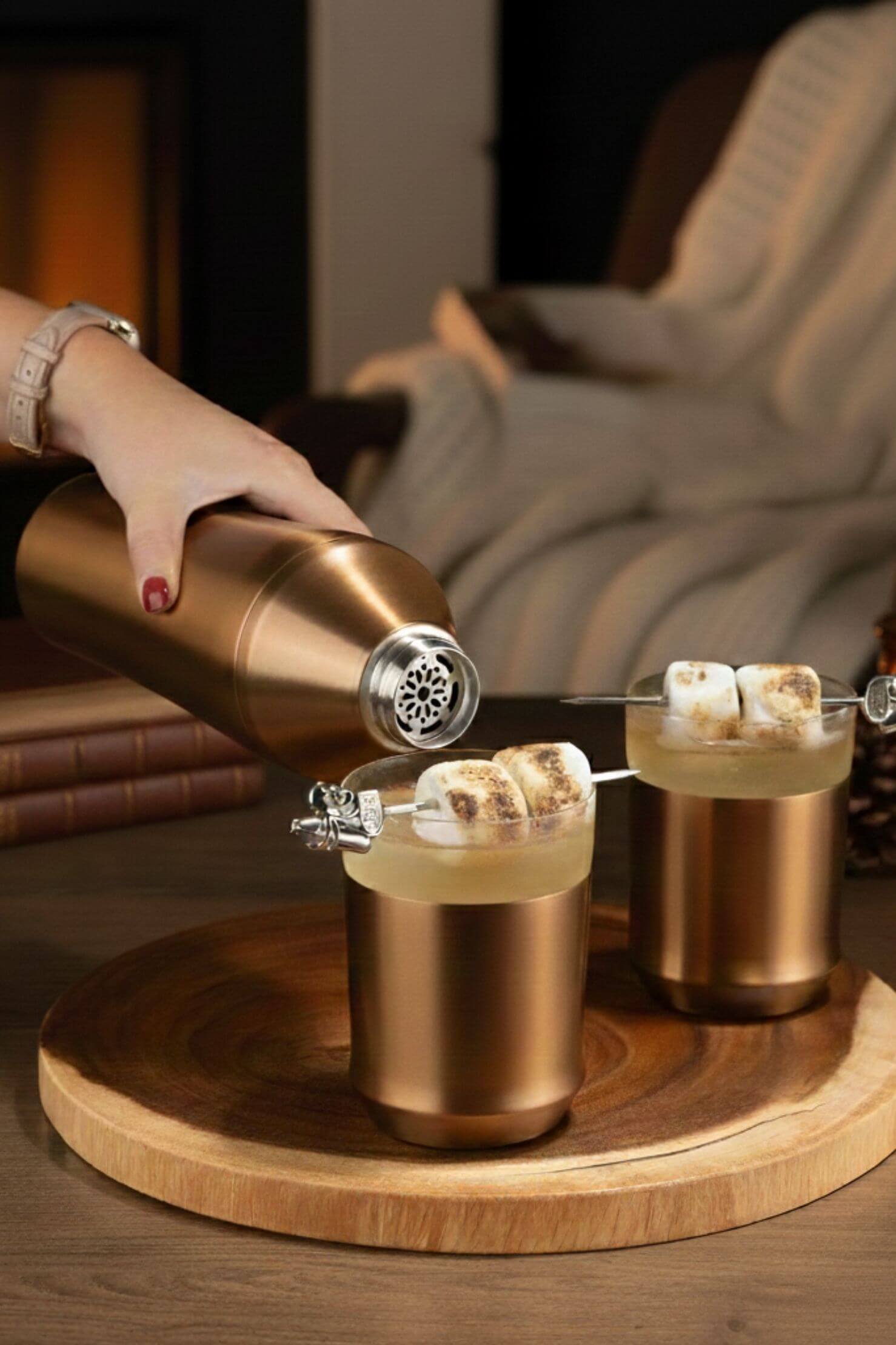 Person pouring a drink into two copper cups with marshmallows on a wooden tray.