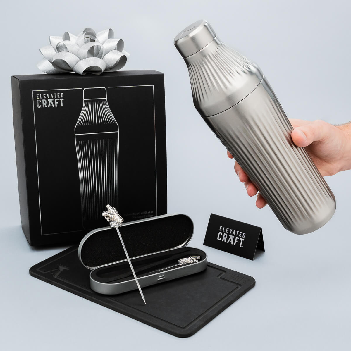 Silver cocktail shaker with packaging and tools on a light gray background