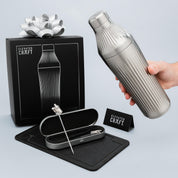 Silver cocktail shaker with packaging and tools on a light gray background