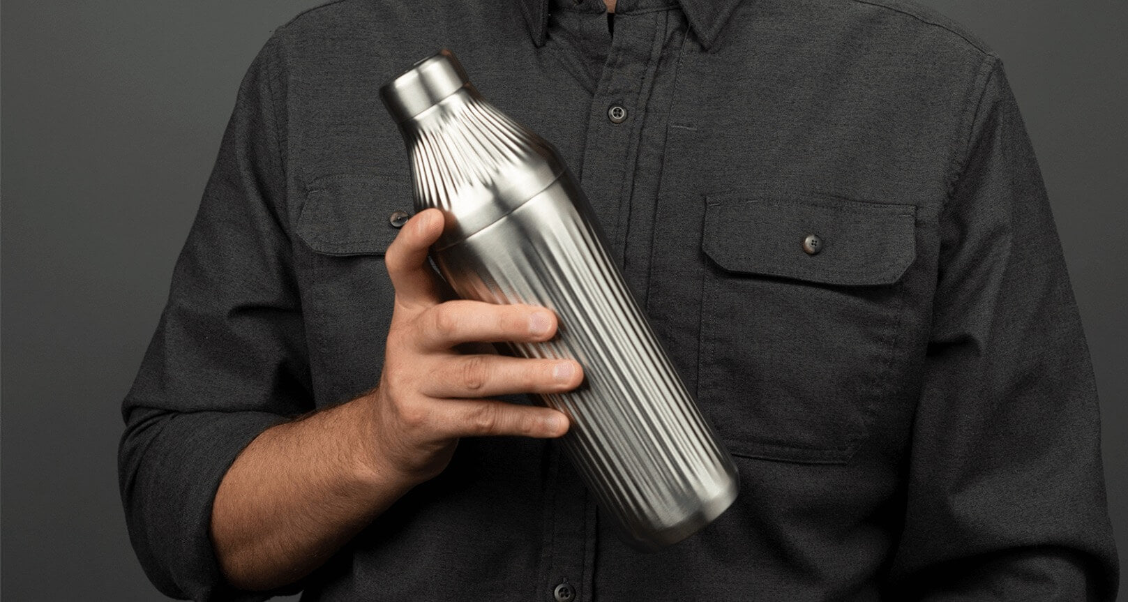 Man holding the Stainless Steel Elevated Craft Diamond Edition Hybrid Cocktail Shaker in front of his grey button up shirt  standing in front of grey background.  