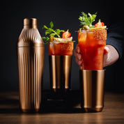 Two Bloody Mary cocktails with garnishes and a bronze shaker on a dark background