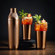 Two Bloody Mary cocktails with garnishes next to a bronze shaker on a dark background