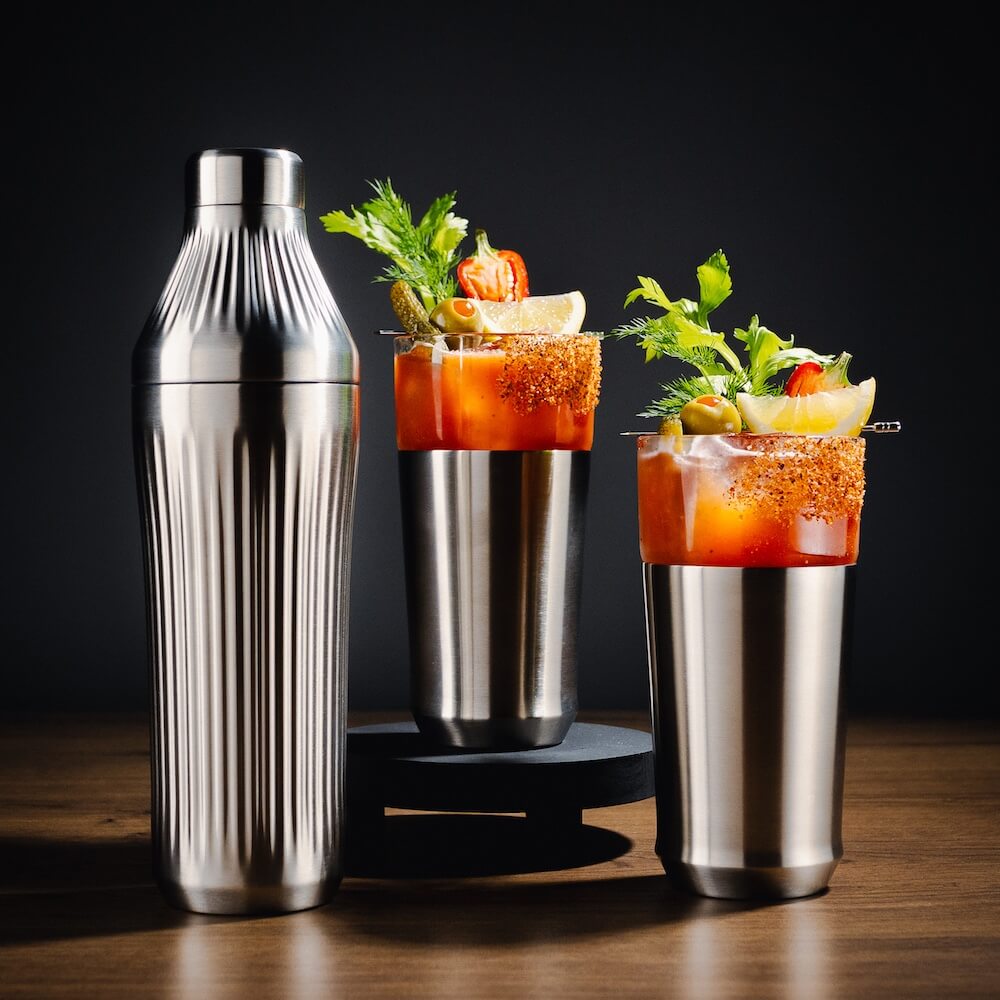 Two Bloody Mary cocktails with garnishes and a cocktail shaker on a wooden surface.