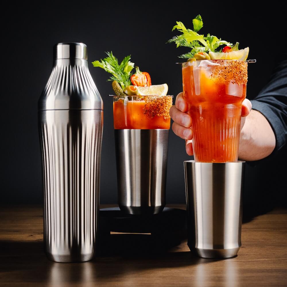 Person holding a Bloody Mary cocktail with a shaker and glasses on a wooden surface.
