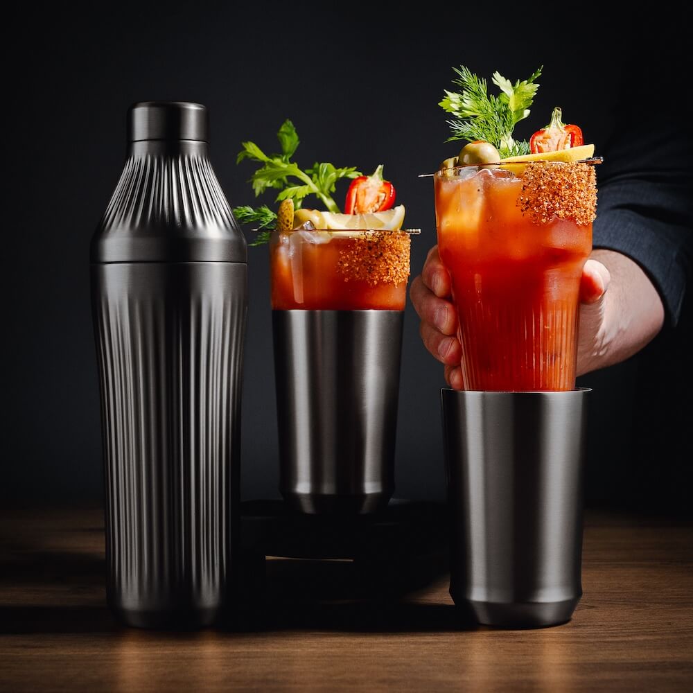 Two Bloody Mary cocktails with garnishes and a shaker on a wooden surface.