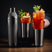 Two Bloody Mary cocktails with garnishes and a shaker on a wooden surface.