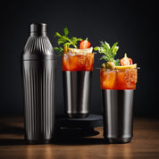 Two Bloody Mary cocktails with garnishes next to a cocktail shaker on a dark background