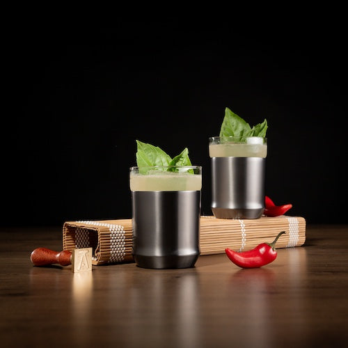 Two cocktails with basil leaves on a wooden surface with red peppers and a pepper grinder.