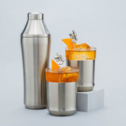 Two cocktail glasses with orange drinks and a metal shaker on a light gray background