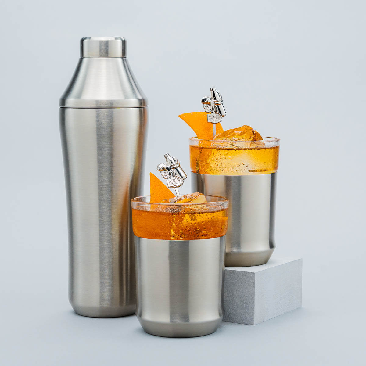 Two cocktail glasses with orange drinks and a metal shaker on a light gray background