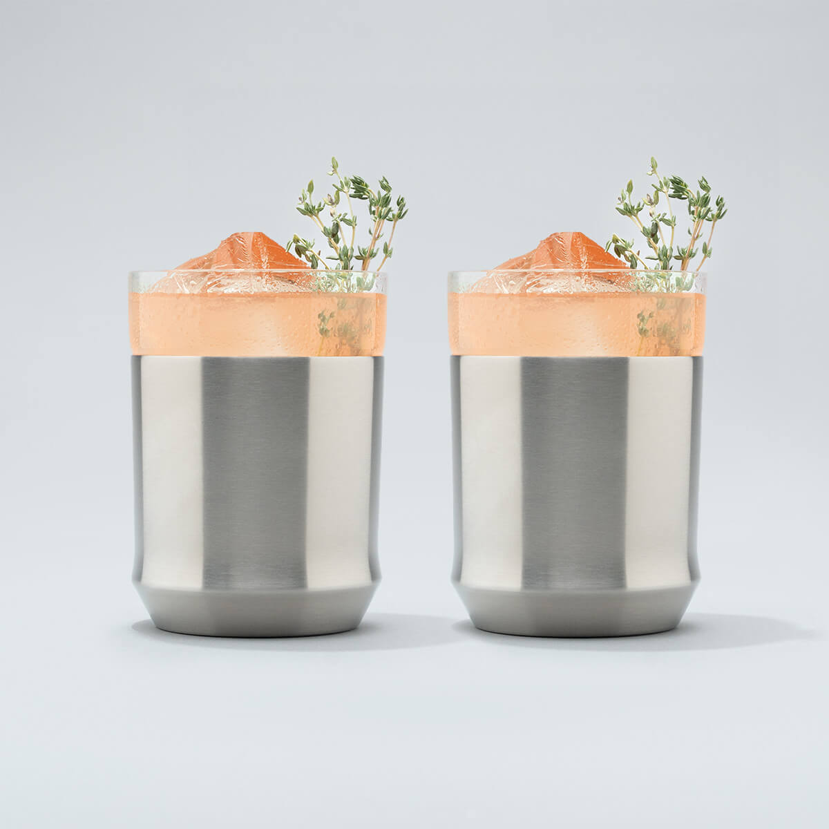 Two Stainless steel Elevated Craft Hybrid Cocktail Glasses filled with bright peach cocktails with herb garnishes
