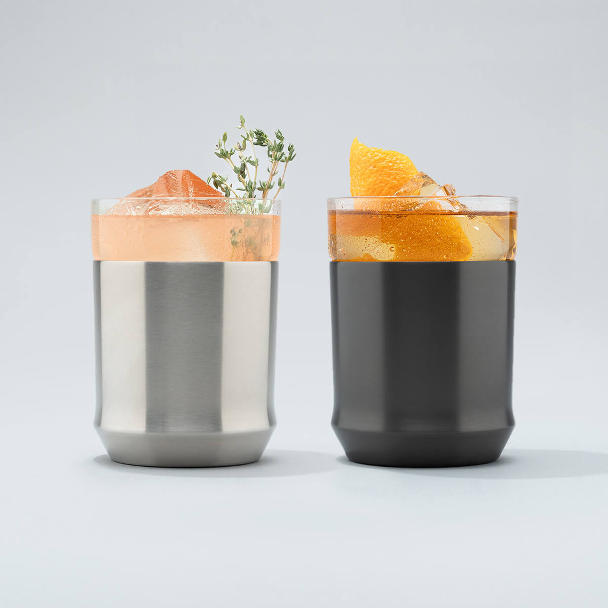 Stainless steel Elevated Craft Hybrid Cocktail Glass filled with bright peach cocktail with an herb garnish sitting next to a gunmetal black hybrid cocktail glass filled with amber cocktail large ice cubes and an orange peel garnish