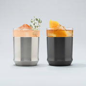 Stainless steel Elevated Craft Hybrid Cocktail Glass filled with bright peach cocktail with an herb garnish sitting next to a gunmetal black hybrid cocktail glass filled with amber cocktail large ice cubes and an orange peel garnish
