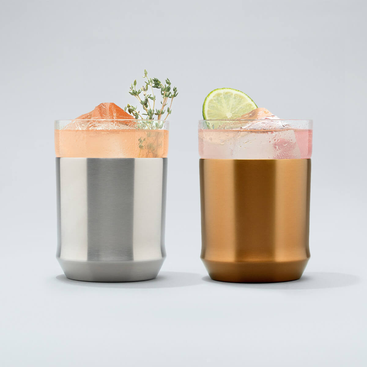 Elevated Craft hybrid cocktail glass in stainless steel finish filled with bright peach cocktail large ice cube and an herb garnish sitting next to a brushed copper hybrid cocktail glass filled with a bright pink cocktail large ice cube and a lime wheel garnish