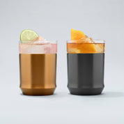 Copper finish Hybrid Cocktail Glass filled with bright pink cocktail garnished with a lime wheel sitting next to a gunmetal black finish hybrid cocktail glass filled with an amber cocktail garnished with an orange peel
