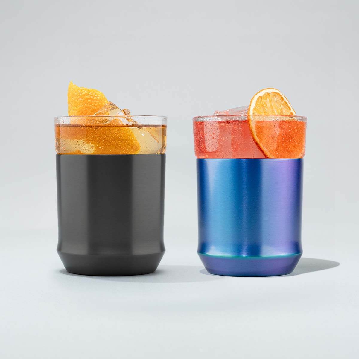 Two hybrid tumblers, one gunmetal color, the other is blue iridescent with drinks and garnishes on a light gray background