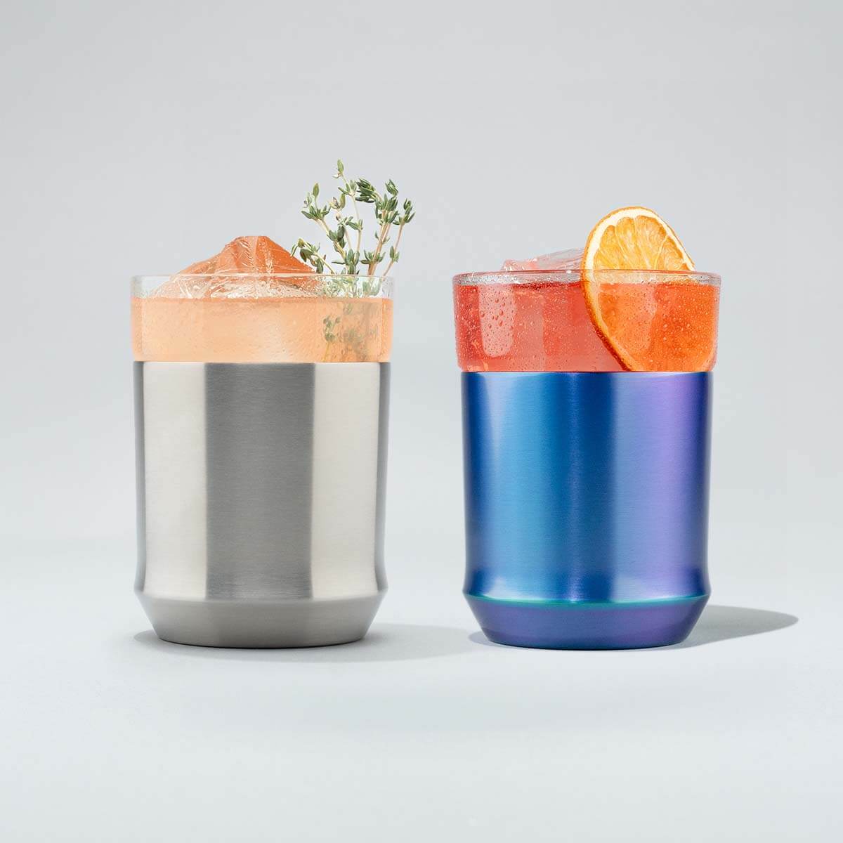 Two hybrid tumblers, one stainless steel, the other is blue Iridescent with drinks and garnishes on a light gray background