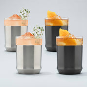 Two Stainless steel Elevated Craft Hybrid Cocktail Glasses filled with bright peach cocktails with herb garnishes sitting next to two gunmetal black hybrid cocktail glasses filled with amber cocktails large ice cubes and orange peel garnishes