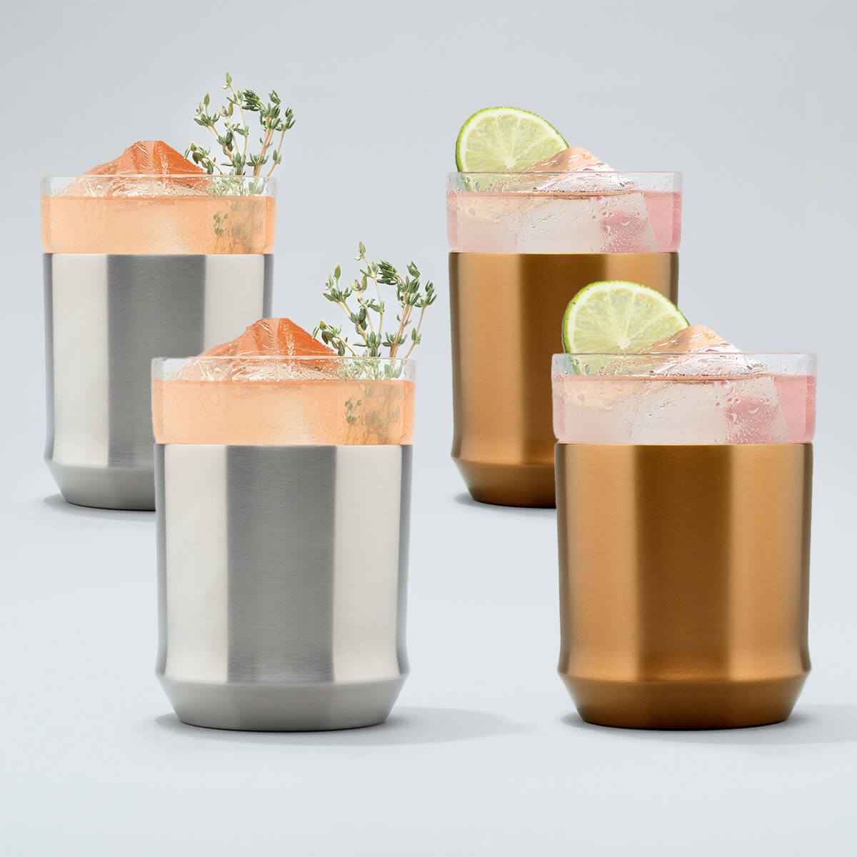 Two Elevated Craft hybrid cocktail glasses in stainless steel finish filled with bright peach cocktails large ice cubes and herb garnishes sitting next to two brushed copper hybrid cocktail glasses filled with bright pink cocktails large ice cubes and lime wheel garnishes