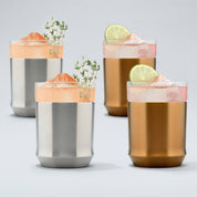 Two Elevated Craft hybrid cocktail glasses in stainless steel finish filled with bright peach cocktails large ice cubes and herb garnishes sitting next to two brushed copper hybrid cocktail glasses filled with bright pink cocktails large ice cubes and lime wheel garnishes
