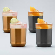 Two Copper finish Hybrid Cocktail Glasses filled with bright pink cocktails garnished with lime wheels sitting next to two gunmetal black finish hybrid cocktail glasses filled with amber cocktails garnished with orange peels