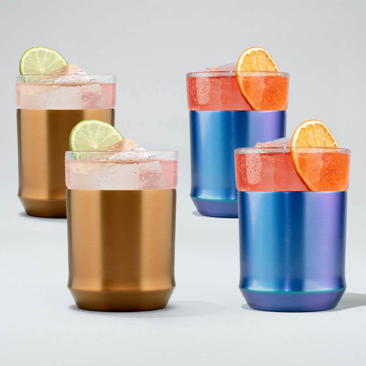 Four metallic tumblers filled with colorful drinks and fruit slices on a light gray background