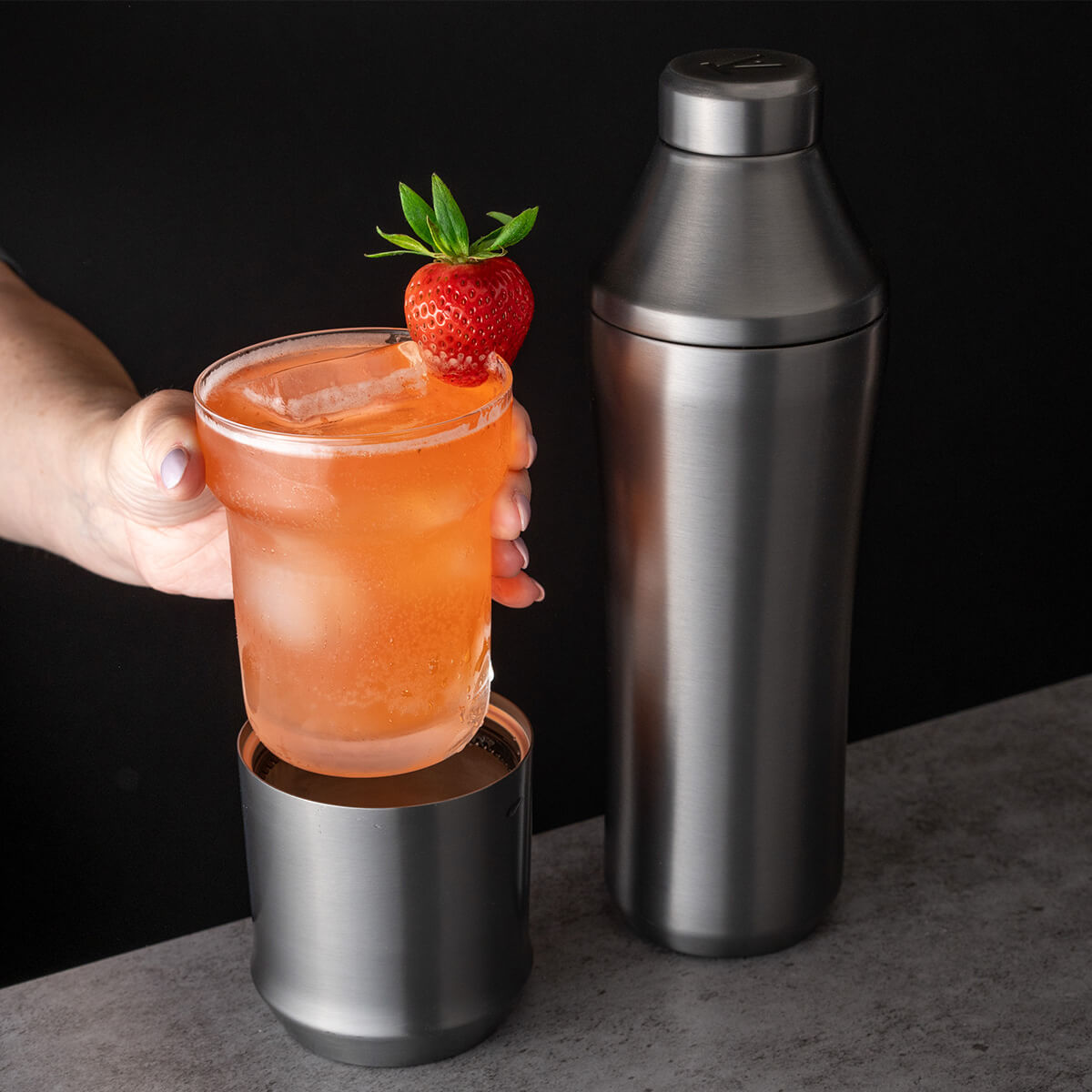 Hand holding glass insert of Elevated Craft Gunmetal Black Hybrid Cocktail Glass filled with peach colored cocktail topped with a strawberry next to the Hybrid Cocktail Shaker