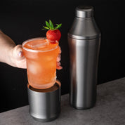 Hand holding glass insert of Elevated Craft Gunmetal Black Hybrid Cocktail Glass filled with peach colored cocktail topped with a strawberry next to the Hybrid Cocktail Shaker