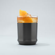 The Elevated Craft Hybrid Cocktail Glass in gunmetal black finish filled with a cold amber cocktail large ice cubes and orange peel garnish