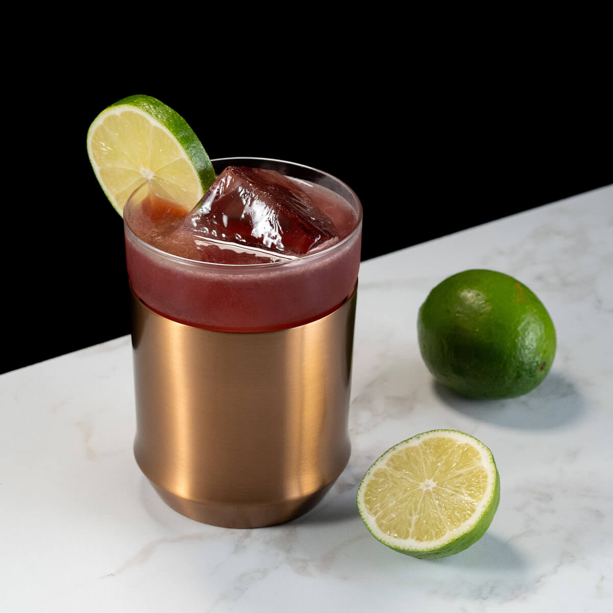 Elevated Craft Brushed Copper Hybrid Cocktail Glass filled with dark red cocktail topped with lime slice next to limes on countertop