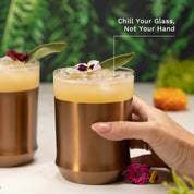 Hand holding the Elevated Craft Brushed Copper Hybrid Cocktail Glass filled with light yellow cocktail topped with edible flower with callout "Chill Your Glass, Not Your Hand"