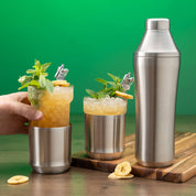 Hand holding glass insert of Elevated Craft Brushed Stainless Hybrid Cocktail Glass filled with yellow colored cocktail topped with mint and banana next to the Hybrid Cocktail Shaker