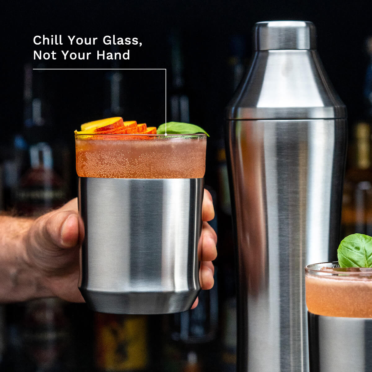 Hand holding the Elevated Craft Brushed Stainless Hybrid Cocktail Glass filled with cocktail topped with peach slices and basil with callout "Chill Your Glass, Not Your Hand"