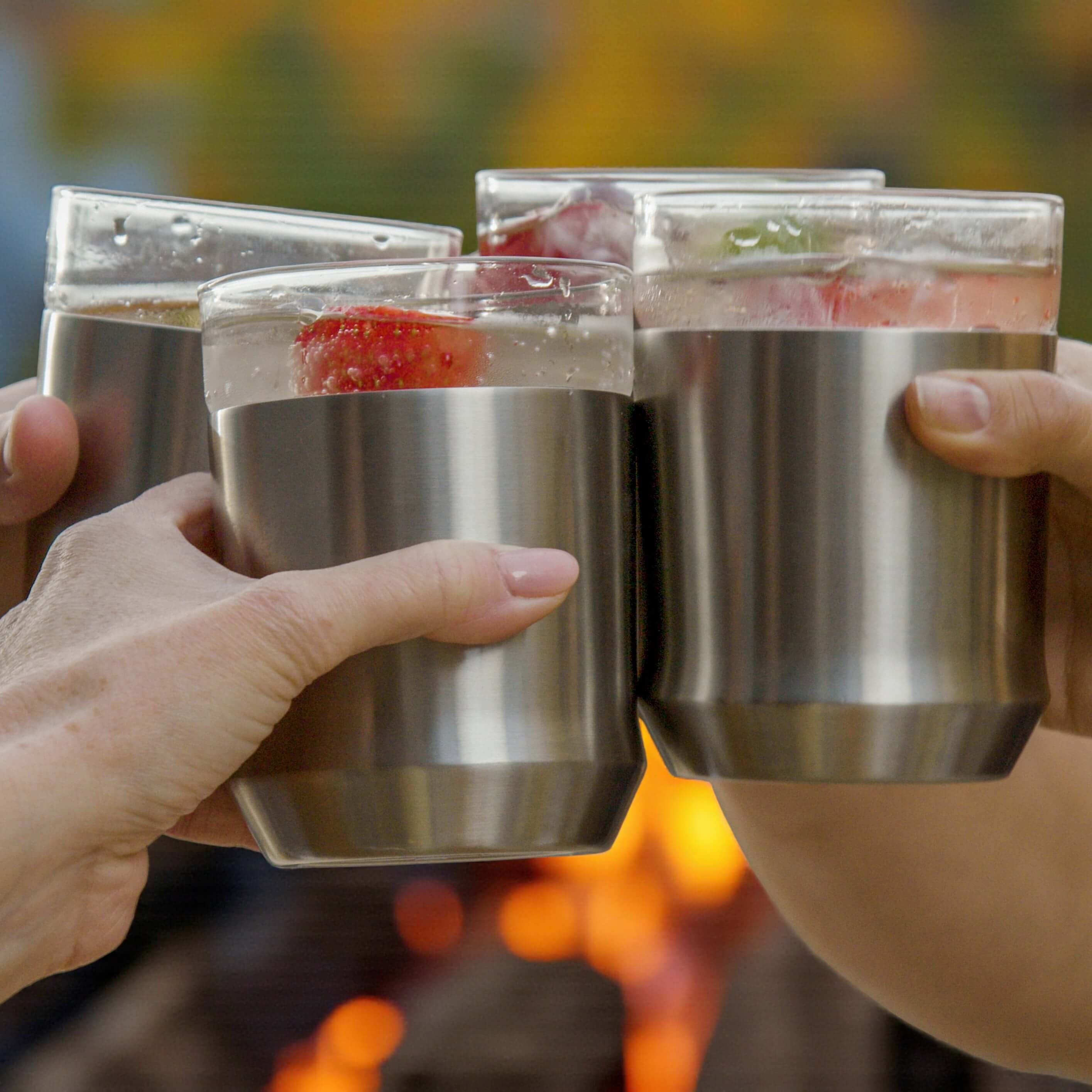 Three hands holding stainless steel tumblers with a blurred background of a campfire.