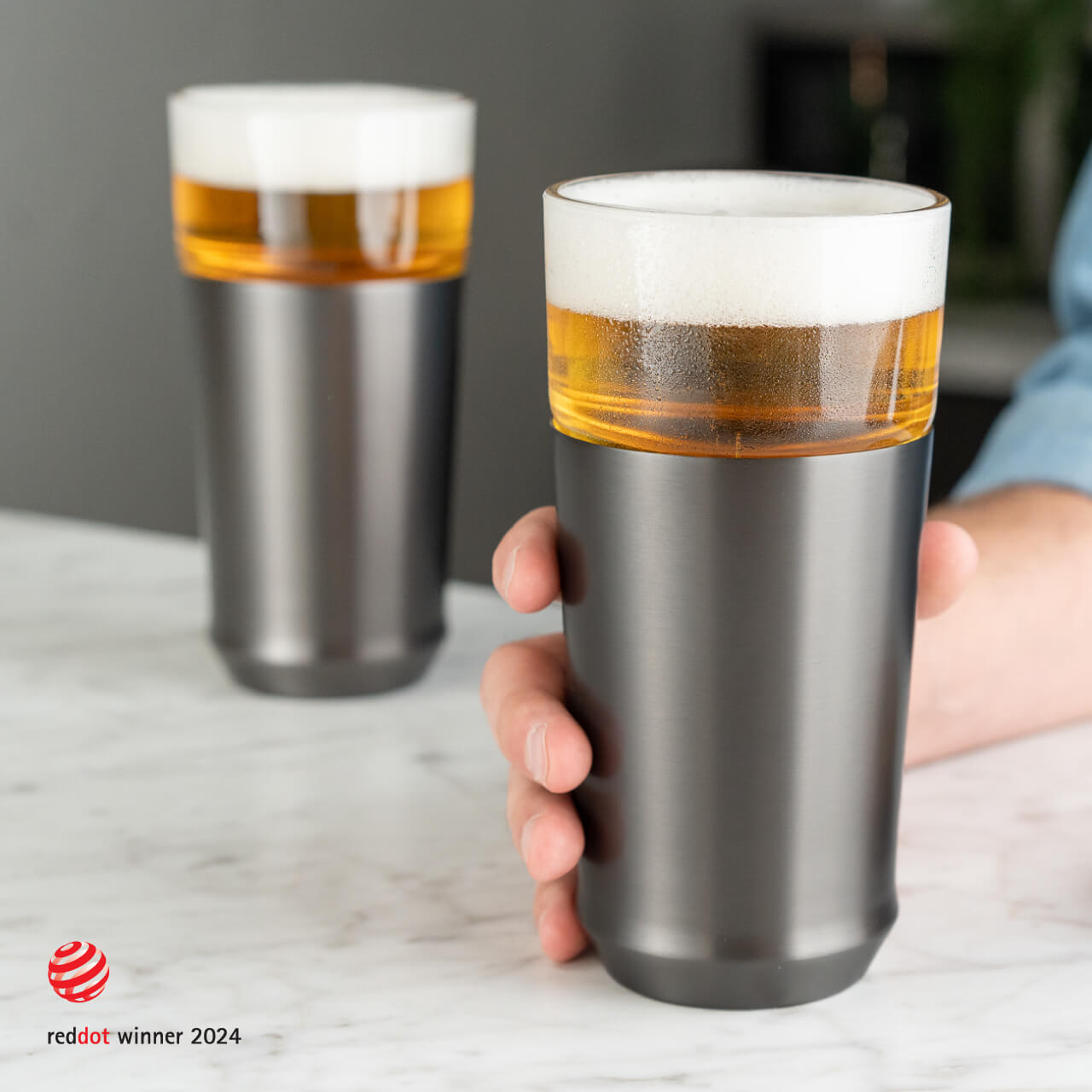 A hand holding the Elevated Craft hybrid pint glass in gunmetal black full of cold beer in the foreground and another gunmetal black hybrid pint glass full of beer in the background  with the 2024 red dot design award logo overlaid in the bottom left corner