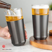 Person pouring cold beer from a bottle into the Elevated Craft Hybrid Pint Glass in gunmetal black finish next to another stainless hybrid pint glass full of cold beer with the 2024 red dot design award logo overlaid in the bottom left corner