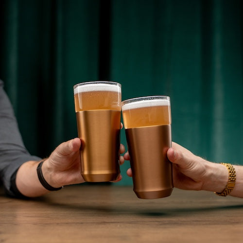 Two hands clinking copper-colored beer glasses against a green curtain background