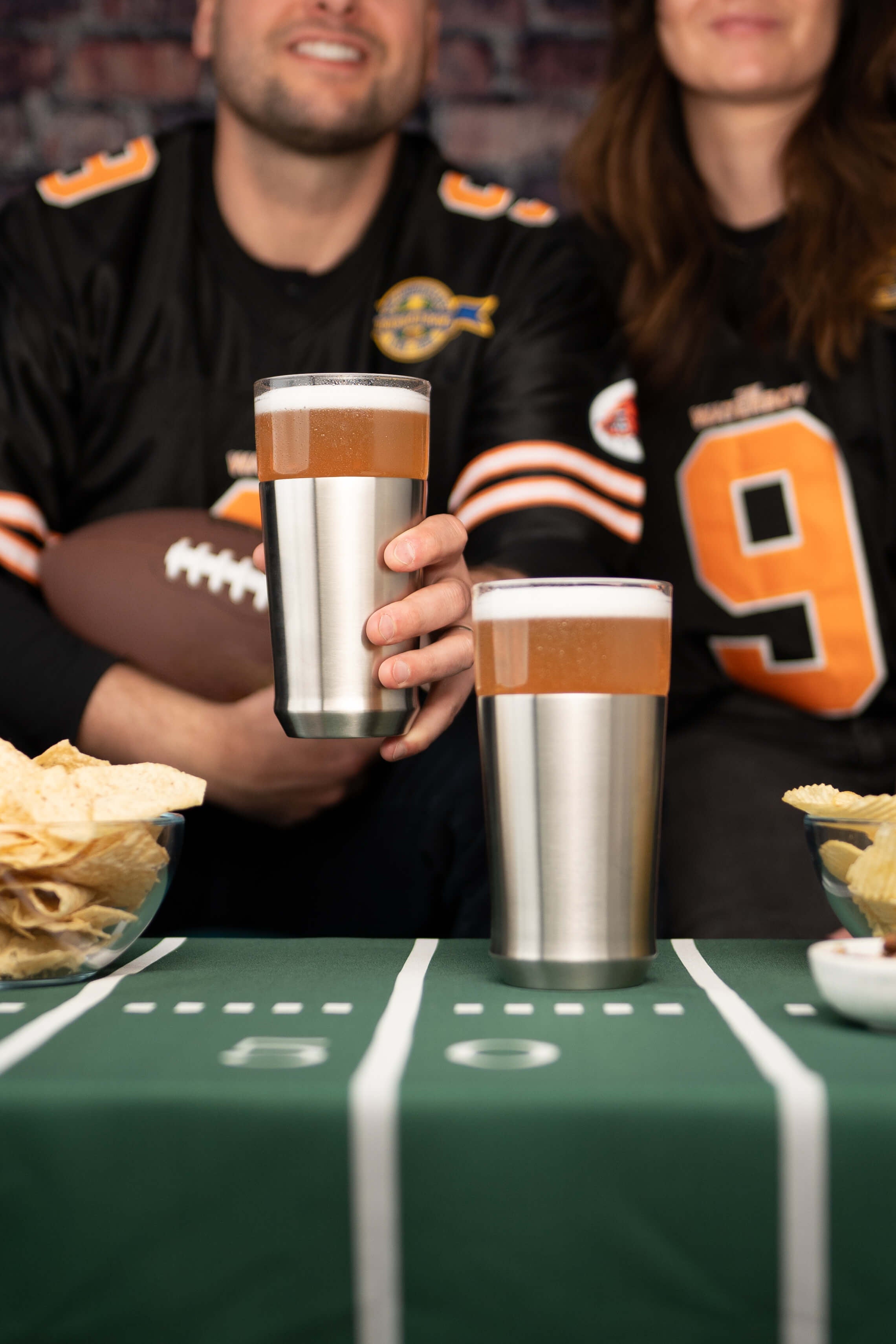 Two people in sports jerseys with a football, holding insulated cups on a table with a sports theme.