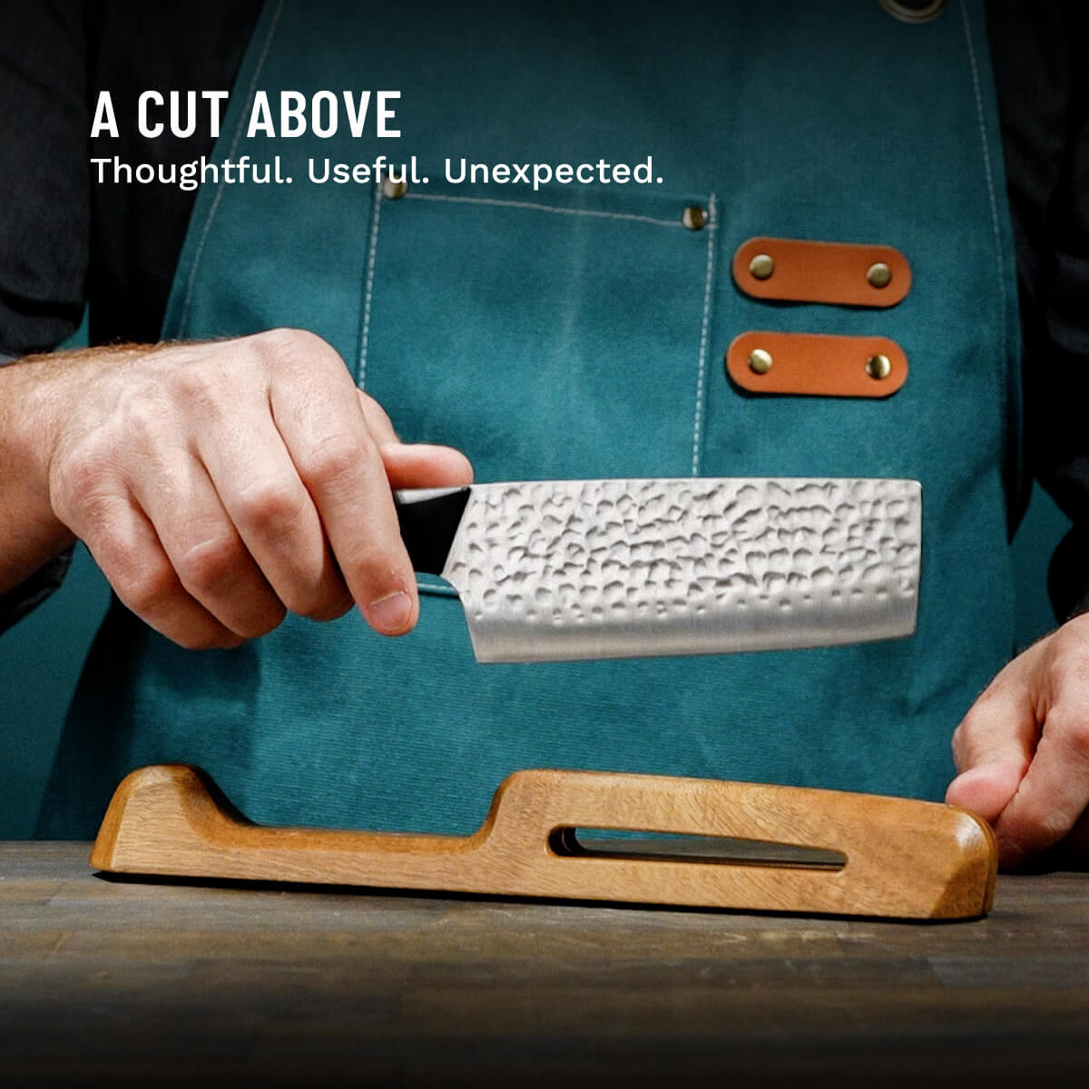 Person holding a knife with a black handle, wearing a blue apron, on a wooden surface.