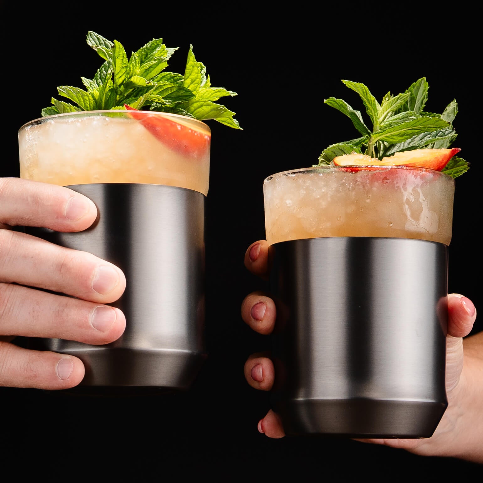 Two hands holding stainless steel cocktail glasses filled with a pale yellow drink, garnished with mint leaves and a slice of citrus, against a solid black background.