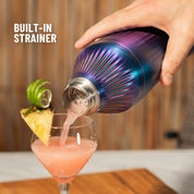 Colorful cocktail shaker being used to pour a pink drink into a glass with a pineapple slice, on a wooden surface.